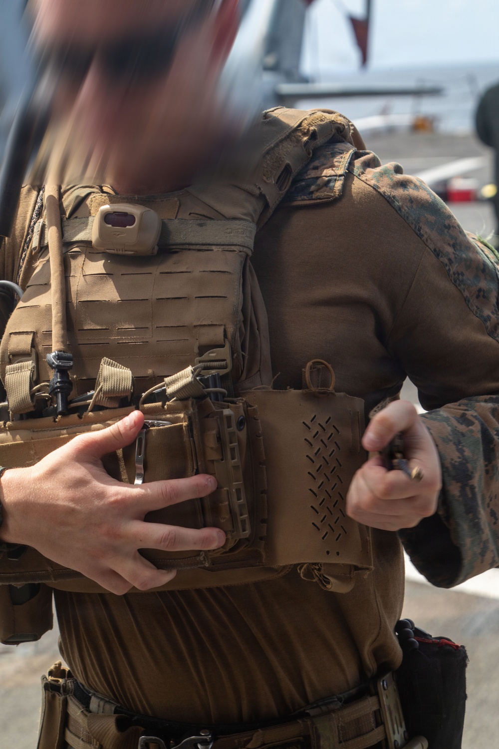 A military personnel wearing USMC MARPAT digital camouflage uniform and a coyote/tan plate carrier vest is handling what appears to be ammunition or a small object. The individual is on what appears to be a ship's flight deck, with aircraft visible in the background. The image is partially obscured by motion blur in the upper left area.