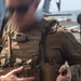 A military personnel wearing USMC MARPAT digital camouflage uniform and a coyote/tan plate carrier vest is handling what appears to be ammunition or a small object. The individual is on what appears to be a ship's flight deck, with aircraft visible in the background. The image is partially obscured by motion blur in the upper left area.