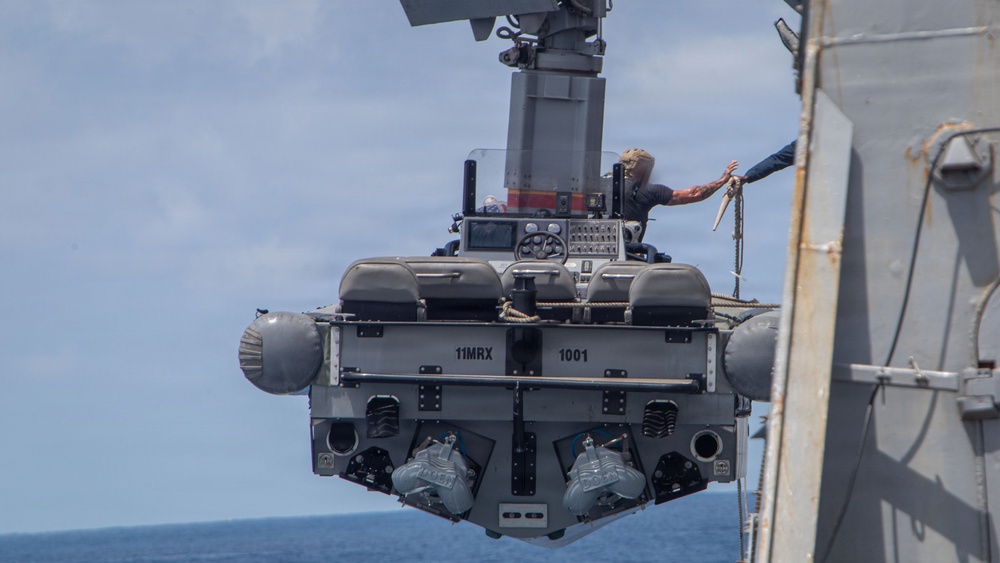 A rigid-hull inflatable boat (RHIB) or small craft designated '11MRX 1001' is being hoisted by a crane davit system alongside a naval vessel. At least one crew member is visible aboard the suspended craft, appearing to coordinate with personnel on the ship's deck. The operation appears to be a boat recovery or launch evolution conducted at sea.