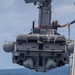 A rigid-hull inflatable boat (RHIB) or small craft designated '11MRX 1001' is being hoisted by a crane davit system alongside a naval vessel. At least one crew member is visible aboard the suspended craft, appearing to coordinate with personnel on the ship's deck. The operation appears to be a boat recovery or launch evolution conducted at sea.