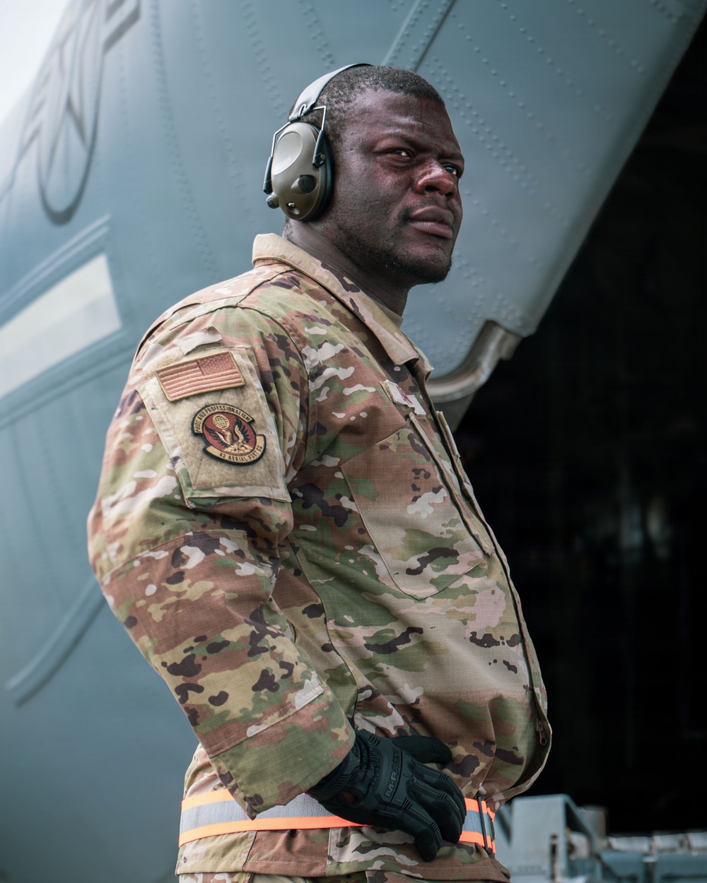 A U.S. Air Force service member stands in front of a large military aircraft, wearing OCP (Operational Camouflage Pattern) uniform with a U.S. flag patch and a unit patch on the left sleeve. The individual is equipped with hearing protection earmuffs, tactical gloves, and a high-visibility orange safety belt, indicating active flight line or aircraft ground operations duties. The portrait-style photograph is taken from a low angle, giving the subject a commanding presence.