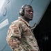 A U.S. Air Force service member stands in front of a large military aircraft, wearing OCP (Operational Camouflage Pattern) uniform with a U.S. flag patch and a unit patch on the left sleeve. The individual is equipped with hearing protection earmuffs, tactical gloves, and a high-visibility orange safety belt, indicating active flight line or aircraft ground operations duties. The portrait-style photograph is taken from a low angle, giving the subject a commanding presence.