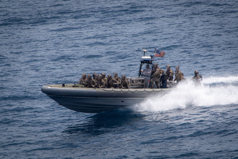 A rigid-hull inflatable boat (RHIB) is underway at high speed on open blue water, carrying multiple personnel in military tactical gear. An American flag is visible mounted on the center console mast. The boat is producing a significant wake, indicating high-speed travel.