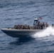 A rigid-hull inflatable boat (RHIB) is underway at high speed on open blue water, carrying multiple personnel in military tactical gear. An American flag is visible mounted on the center console mast. The boat is producing a significant wake, indicating high-speed travel.