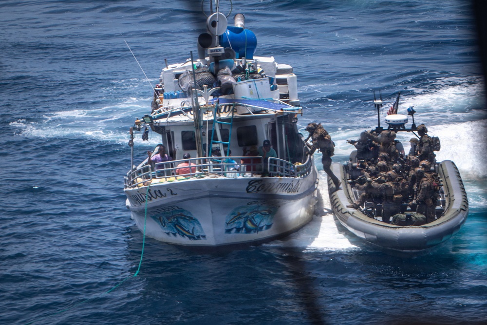 An aerial photograph shows a military or law enforcement interdiction operation at sea. Armed personnel in tactical gear aboard a rigid inflatable boat (RIB) are approaching and boarding a white civilian fishing vessel named 'Conquistador 2'. The operation appears to be a maritime law enforcement or counter-narcotics boarding action in open ocean waters.