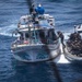 An aerial photograph shows a military or law enforcement interdiction operation at sea. Armed personnel in tactical gear aboard a rigid inflatable boat (RIB) are approaching and boarding a white civilian fishing vessel named 'Conquistador 2'. The operation appears to be a maritime law enforcement or counter-narcotics boarding action in open ocean waters.