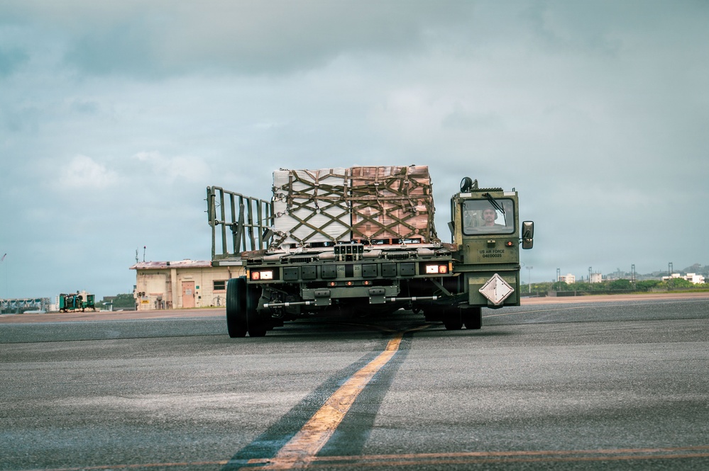 A US Air Force military flatbed truck is transporting a palletized cargo load across an airfield tarmac. The cargo is secured with netting on what appears to be a standard military air cargo pallet. The setting appears to be a military airfield, likely in a tropical or coastal region based on the vegetation and sky conditions.