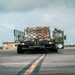 A US Air Force military flatbed truck is transporting a palletized cargo load across an airfield tarmac. The cargo is secured with netting on what appears to be a standard military air cargo pallet. The setting appears to be a military airfield, likely in a tropical or coastal region based on the vegetation and sky conditions.