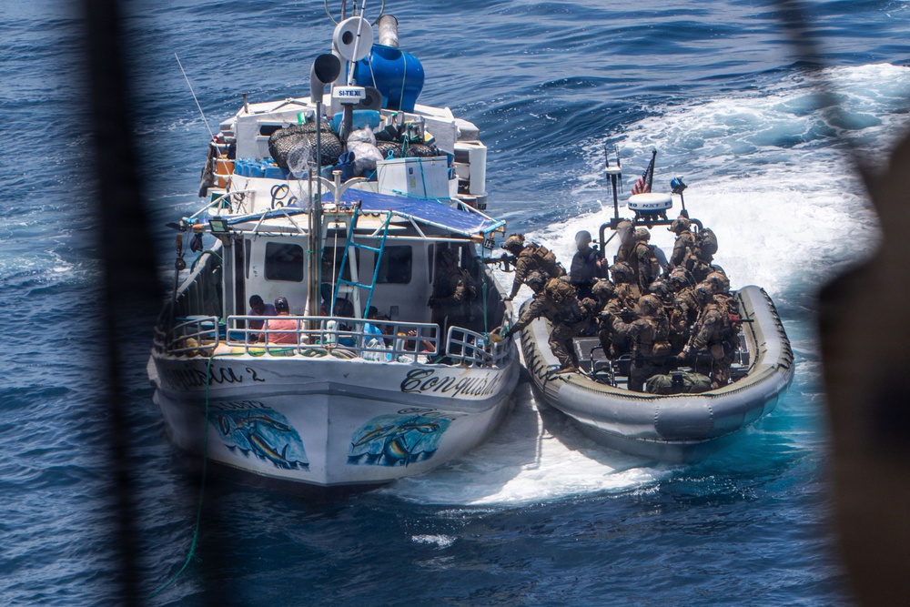 A military or law enforcement interdiction operation is underway at sea, with a rigid inflatable boat (RIB) carrying armed personnel in tactical gear approaching a civilian-style fishing vessel named 'Conquista 2'. The armed personnel appear to be boarding or preparing to board the fishing vessel. An American flag is visible on the RIB, suggesting U.S. military or Coast Guard involvement.