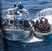 A military or law enforcement interdiction operation is underway at sea, with a rigid inflatable boat (RIB) carrying armed personnel in tactical gear approaching a civilian-style fishing vessel named 'Conquista 2'. The armed personnel appear to be boarding or preparing to board the fishing vessel. An American flag is visible on the RIB, suggesting U.S. military or Coast Guard involvement.