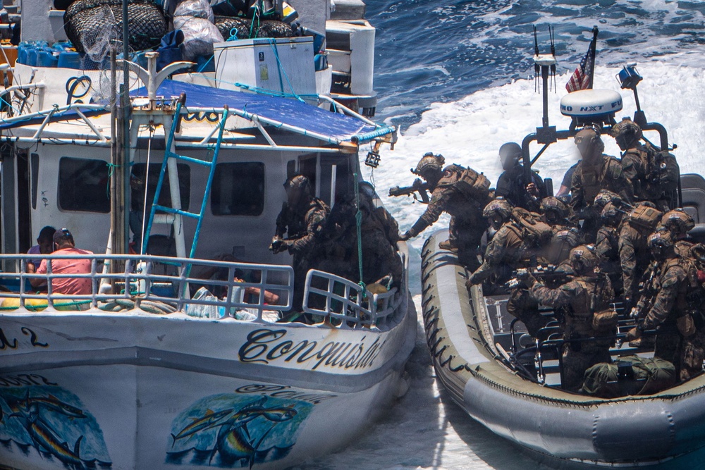The image depicts a maritime law enforcement or military interdiction operation in open ocean waters. Armed personnel in camouflage tactical gear aboard a rigid inflatable boat (RIB) are boarding or approaching a civilian vessel named 'Conquistador.' An American flag is visible on the tactical vessel, suggesting U.S. military or law enforcement involvement.