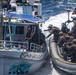 The image depicts a maritime law enforcement or military interdiction operation in open ocean waters. Armed personnel in camouflage tactical gear aboard a rigid inflatable boat (RIB) are boarding or approaching a civilian vessel named 'Conquistador.' An American flag is visible on the tactical vessel, suggesting U.S. military or law enforcement involvement.
