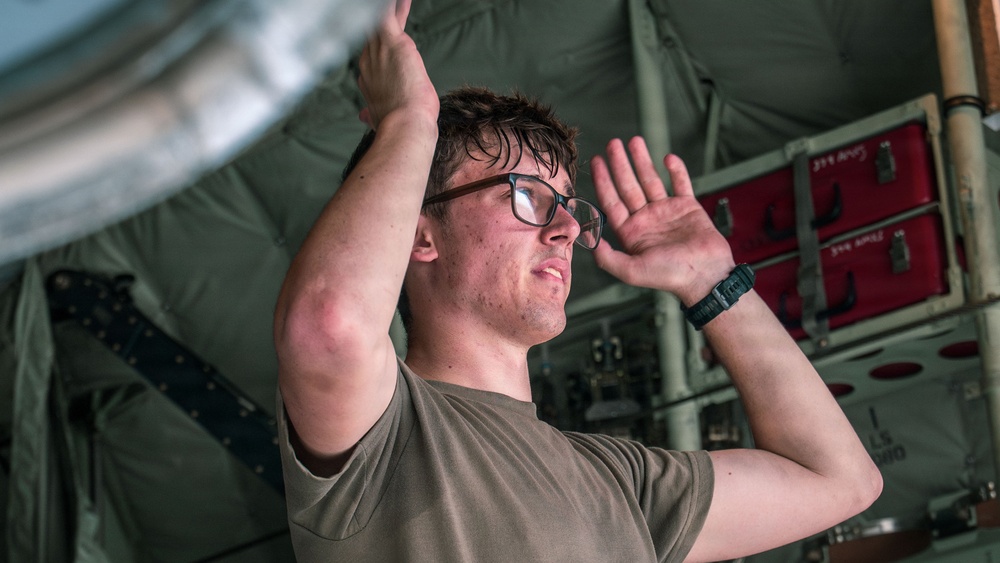 A young male technician or military personnel wearing an olive/tan t-shirt and glasses is working overhead, reaching up with both arms toward a metallic component. The image is taken from a low angle looking upward, showing the interior of what appears to be a military aircraft or vehicle. Red equipment cases and olive-green canvas or paneling are visible in the background.