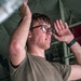 A young male technician or military personnel wearing an olive/tan t-shirt and glasses is working overhead, reaching up with both arms toward a metallic component. The image is taken from a low angle looking upward, showing the interior of what appears to be a military aircraft or vehicle. Red equipment cases and olive-green canvas or paneling are visible in the background.