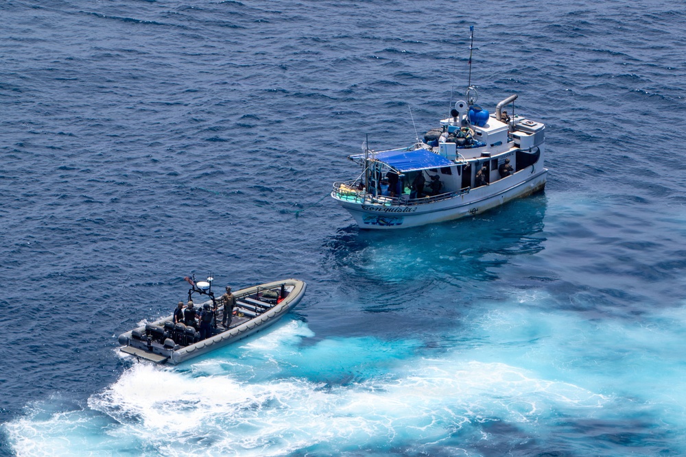 An aerial photograph shows a rigid inflatable boat (RIB) with personnel approaching a larger civilian-style fishing or patrol vessel on open blue ocean waters. The smaller tactical craft appears to be conducting an intercept or boarding operation. The larger vessel has a blue tarp canopy and multiple people visible on deck.