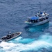 An aerial photograph shows a rigid inflatable boat (RIB) with personnel approaching a larger civilian-style fishing or patrol vessel on open blue ocean waters. The smaller tactical craft appears to be conducting an intercept or boarding operation. The larger vessel has a blue tarp canopy and multiple people visible on deck.