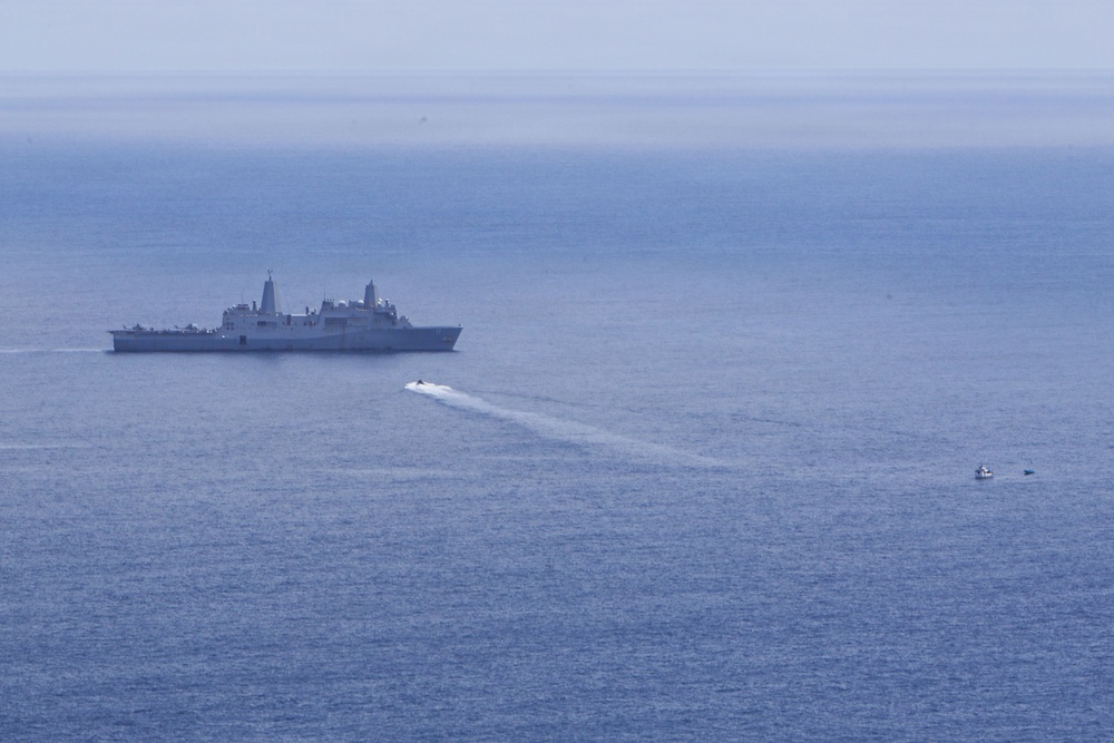 An aerial or elevated view of open ocean showing a large gray naval warship, likely an amphibious transport dock (LPD-class), stationary or slowly moving in the distance. A smaller fast-moving craft is visible creating a white wake trail as it moves away from the larger vessel toward the right side of the frame. Two additional small watercraft are visible at the far right of the image.