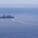 An aerial or elevated view of open ocean showing a large gray naval warship, likely an amphibious transport dock (LPD-class), stationary or slowly moving in the distance. A smaller fast-moving craft is visible creating a white wake trail as it moves away from the larger vessel toward the right side of the frame. Two additional small watercraft are visible at the far right of the image.