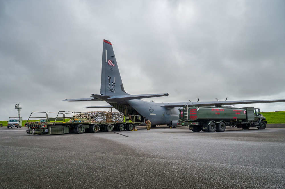 A U.S. Air Force C-130 Hercules transport aircraft (tail code YJ, 5 AF, serial 833) is being serviced on a flight line ramp under overcast skies. Ground crews are simultaneously loading cargo pallets via the rear ramp and refueling the aircraft using a military fuel tanker truck. The scene depicts a typical forward operating base or expeditionary airfield logistics operation.
