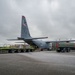 A U.S. Air Force C-130 Hercules transport aircraft (tail code YJ, 5 AF, serial 833) is being serviced on a flight line ramp under overcast skies. Ground crews are simultaneously loading cargo pallets via the rear ramp and refueling the aircraft using a military fuel tanker truck. The scene depicts a typical forward operating base or expeditionary airfield logistics operation.