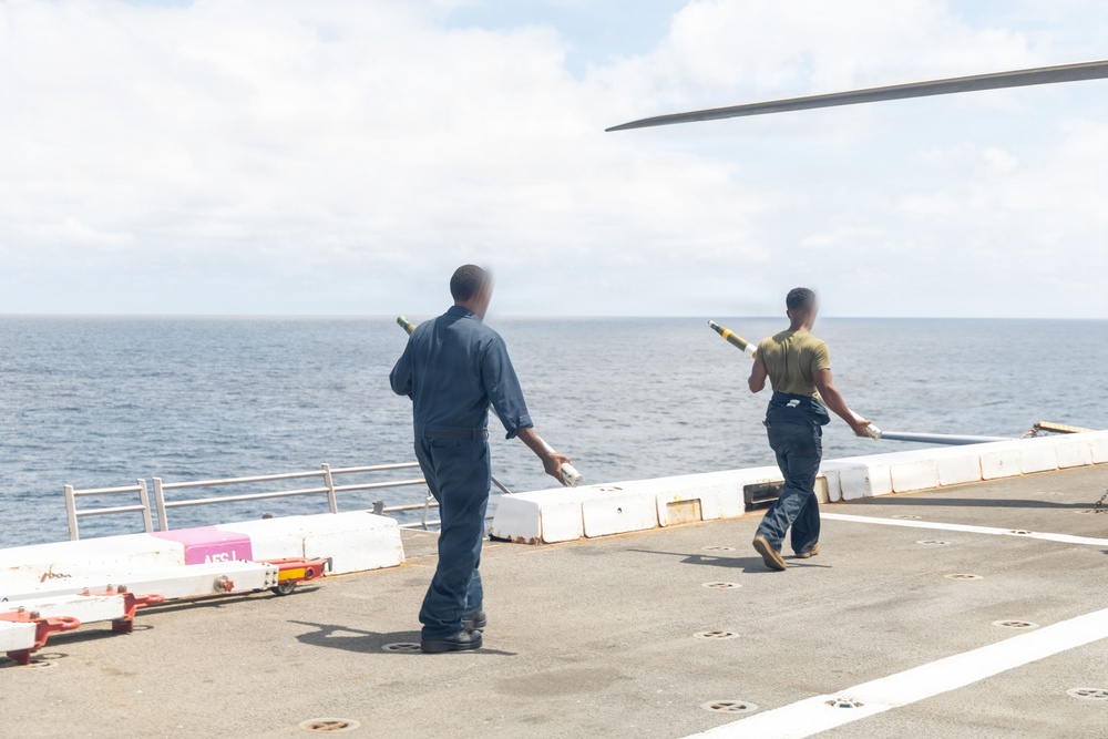 Two military personnel are carrying what appear to be missile or rocket components across a ship's flight deck at sea. They are walking toward the edge of the deck, with the ocean visible in the background. A partial rotor blade or wing is visible in the upper right corner of the frame.