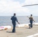 Two military personnel are carrying what appear to be missile or rocket components across a ship's flight deck at sea. They are walking toward the edge of the deck, with the ocean visible in the background. A partial rotor blade or wing is visible in the upper right corner of the frame.