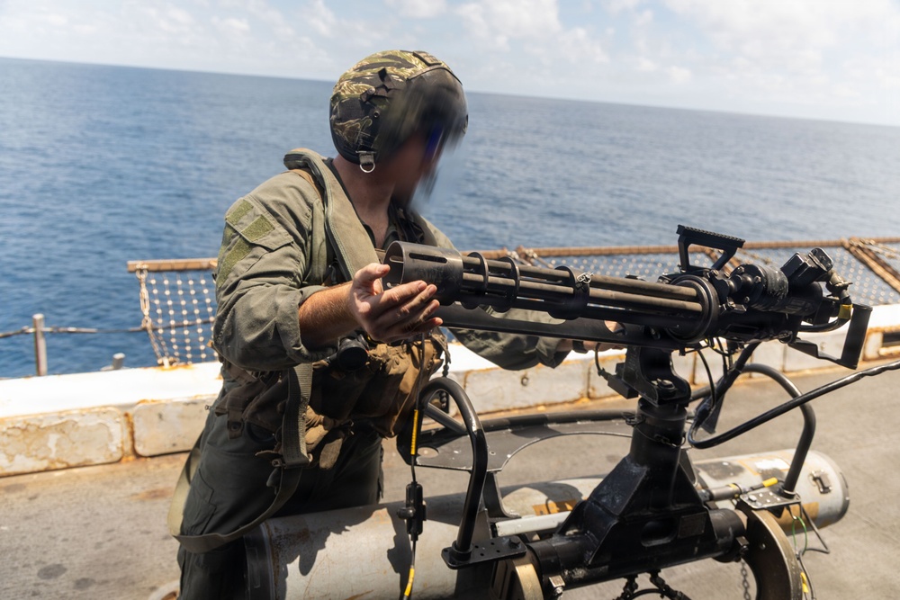 A military service member in camouflage gear is operating or inspecting a mounted multi-barrel rotary weapon system (consistent with an M134 Minigun or similar Gatling-type weapon) on the deck of a naval vessel. The setting is an open ocean environment under clear skies. The person's face is motion-blurred, likely due to camera movement during exposure.