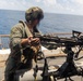 A military service member in camouflage gear is operating or inspecting a mounted multi-barrel rotary weapon system (consistent with an M134 Minigun or similar Gatling-type weapon) on the deck of a naval vessel. The setting is an open ocean environment under clear skies. The person's face is motion-blurred, likely due to camera movement during exposure.