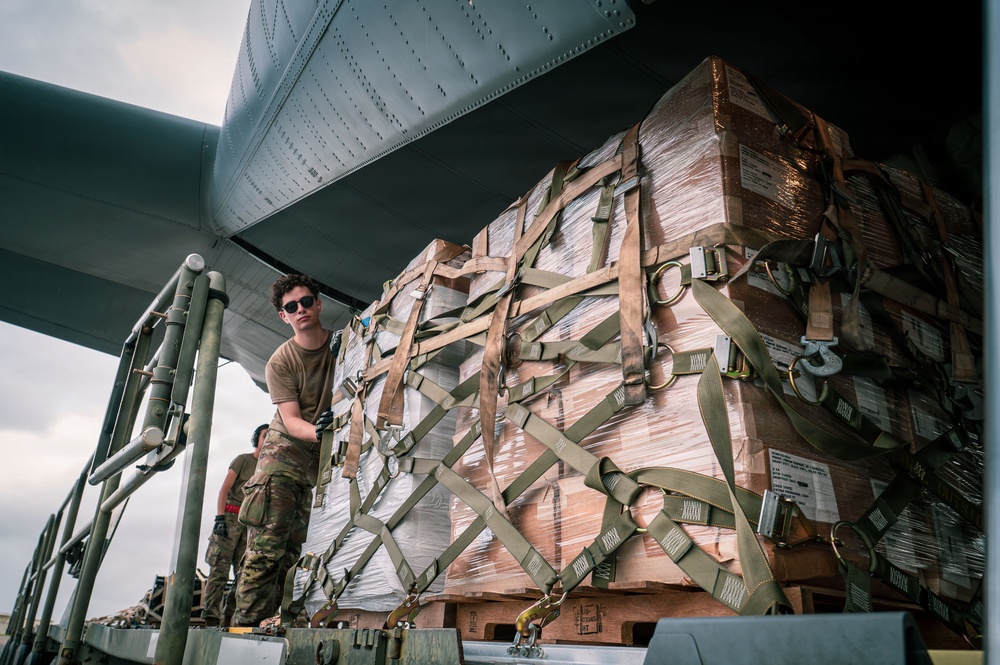 Military personnel are loading or unloading a large palletized cargo shipment from what appears to be a military transport aircraft. The cargo is secured with multiple tan/olive-colored cargo straps and wrapped in clear plastic sheeting over cardboard boxes. At least two service members in camouflage uniforms are visible managing the operation.