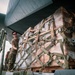 Military personnel are loading or unloading a large palletized cargo shipment from what appears to be a military transport aircraft. The cargo is secured with multiple tan/olive-colored cargo straps and wrapped in clear plastic sheeting over cardboard boxes. At least two service members in camouflage uniforms are visible managing the operation.