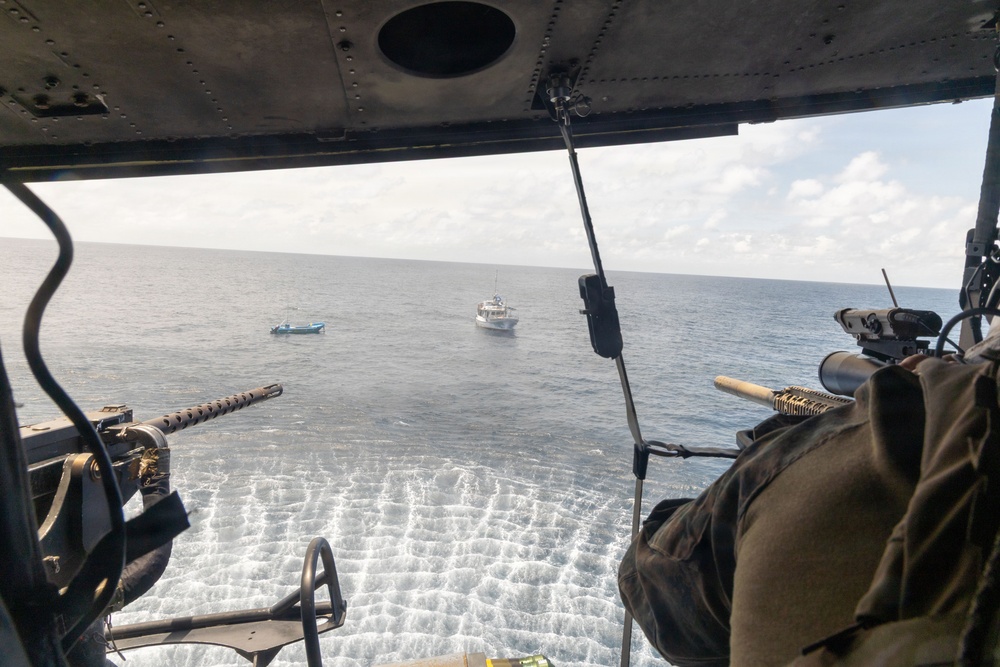 The image is taken from inside a military helicopter looking out over open ocean. Two vessels are visible in the water below — a smaller blue boat and a larger white patrol/coast guard-style vessel. The helicopter appears to be equipped with mounted weapons on both sides of the open door.