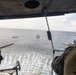The image is taken from inside a military helicopter looking out over open ocean. Two vessels are visible in the water below — a smaller blue boat and a larger white patrol/coast guard-style vessel. The helicopter appears to be equipped with mounted weapons on both sides of the open door.