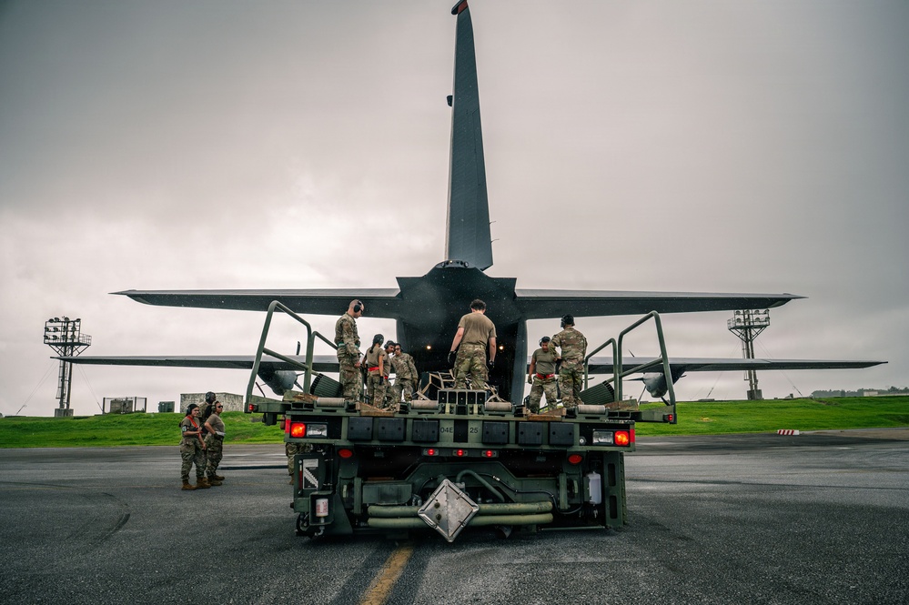 Military personnel in camouflage uniforms are conducting a loading or unloading operation at the rear of a large military transport aircraft, likely a C-130 Hercules, on a wet tarmac under overcast skies. A military flatbed vehicle or loader is positioned directly at the aircraft's open rear ramp, with multiple service members working on and around it. The setting appears to be an active military airfield with lighting towers visible in the background.