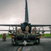 Military personnel in camouflage uniforms are conducting a loading or unloading operation at the rear of a large military transport aircraft, likely a C-130 Hercules, on a wet tarmac under overcast skies. A military flatbed vehicle or loader is positioned directly at the aircraft's open rear ramp, with multiple service members working on and around it. The setting appears to be an active military airfield with lighting towers visible in the background.