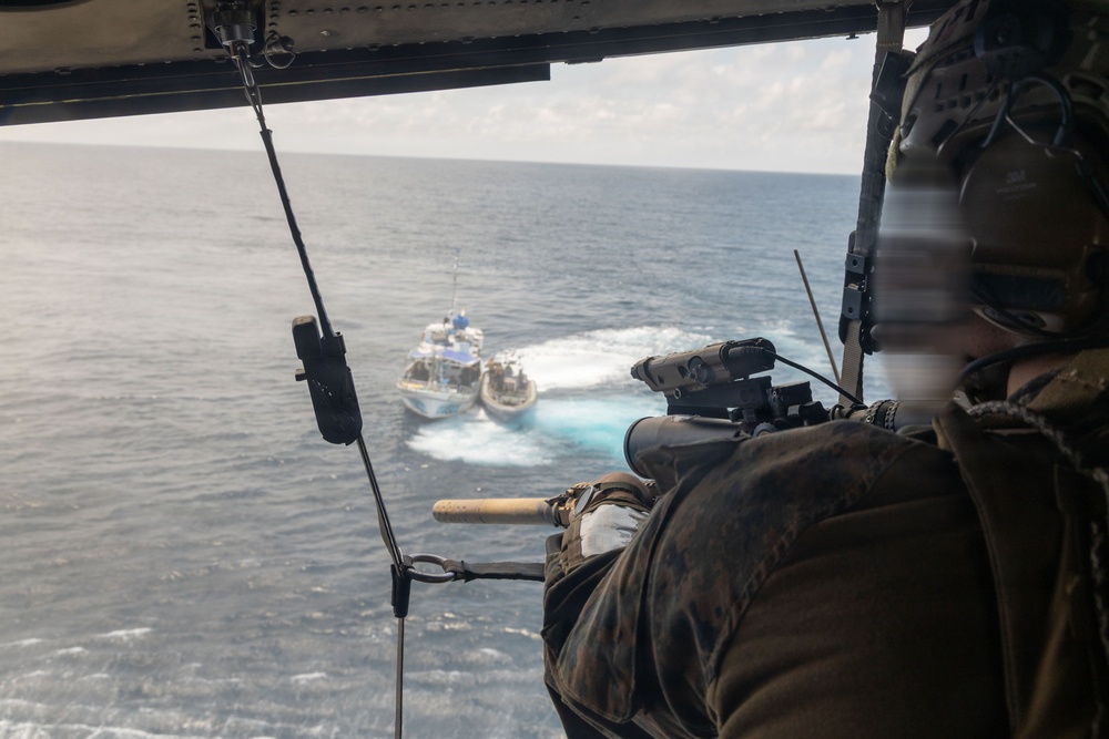 A military or law enforcement personnel is positioned at the open door of a helicopter, aiming a scoped rifle toward a vessel on the open ocean below. The shooter is wearing tactical gear and appears to be observing or targeting a white and blue patrol-style boat that is moving at speed, creating a visible wake. The perspective is from inside the helicopter looking outward and downward toward the water.