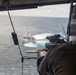 A military or law enforcement personnel is positioned at the open door of a helicopter, aiming a scoped rifle toward a vessel on the open ocean below. The shooter is wearing tactical gear and appears to be observing or targeting a white and blue patrol-style boat that is moving at speed, creating a visible wake. The perspective is from inside the helicopter looking outward and downward toward the water.