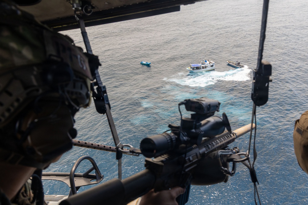 An aerial photograph taken from inside a military helicopter showing a sniper or marksman positioned at the open door with a scoped rifle aimed toward vessels on the ocean below. Three boats are visible on the water, with what appears to be a maritime interdiction or surveillance operation in progress. A second armed personnel member is visible in the background of the helicopter interior.