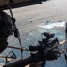 An aerial photograph taken from inside a military helicopter showing a sniper or marksman positioned at the open door with a scoped rifle aimed toward vessels on the ocean below. Three boats are visible on the water, with what appears to be a maritime interdiction or surveillance operation in progress. A second armed personnel member is visible in the background of the helicopter interior.