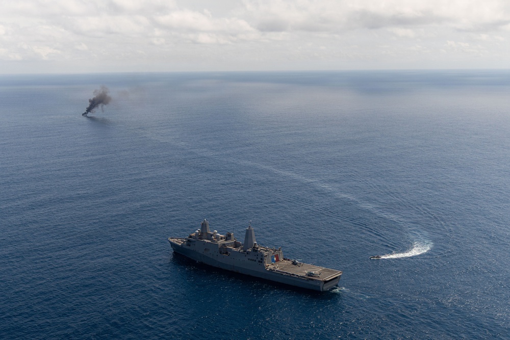 An aerial photograph shows a large gray naval vessel, consistent with a San Antonio-class amphibious transport dock ship, underway in open blue ocean. In the background to the left, a smaller vessel or target is visibly burning and emitting a thick column of black smoke. A small fast-moving craft is visible to the right of the main ship, leaving a white wake.