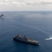 An aerial photograph shows a large gray naval vessel, consistent with a San Antonio-class amphibious transport dock ship, underway in open blue ocean. In the background to the left, a smaller vessel or target is visibly burning and emitting a thick column of black smoke. A small fast-moving craft is visible to the right of the main ship, leaving a white wake.