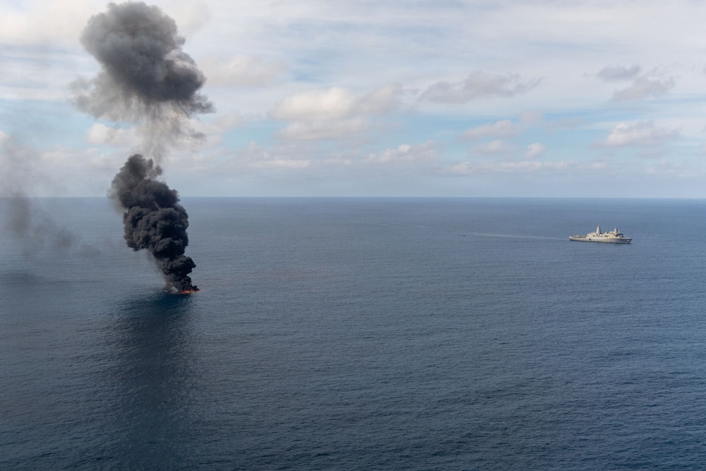 An aerial photograph shows a vessel or object burning on the open ocean, producing a large column of dense black smoke rising into a partly cloudy sky. A naval or military ship is visible in the background to the right, positioned at a safe distance from the fire. The scene appears to depict a maritime incident, possibly a controlled sinking exercise or an engagement at sea.