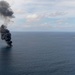 An aerial photograph shows a vessel or object burning on the open ocean, producing a large column of dense black smoke rising into a partly cloudy sky. A naval or military ship is visible in the background to the right, positioned at a safe distance from the fire. The scene appears to depict a maritime incident, possibly a controlled sinking exercise or an engagement at sea.