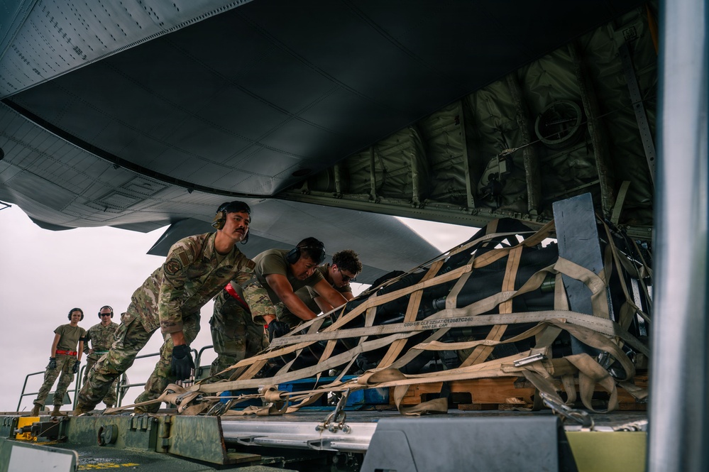 Military personnel in camouflage uniforms are loading or unloading cargo secured with netting and straps onto or from a large military aircraft. The scene appears to take place on a flight line or tarmac, with multiple service members actively handling the netted cargo pallet. The low-angle perspective emphasizes the scale of the aircraft and the physical effort involved in the operation.