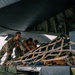 Military personnel in camouflage uniforms are loading or unloading cargo secured with netting and straps onto or from a large military aircraft. The scene appears to take place on a flight line or tarmac, with multiple service members actively handling the netted cargo pallet. The low-angle perspective emphasizes the scale of the aircraft and the physical effort involved in the operation.