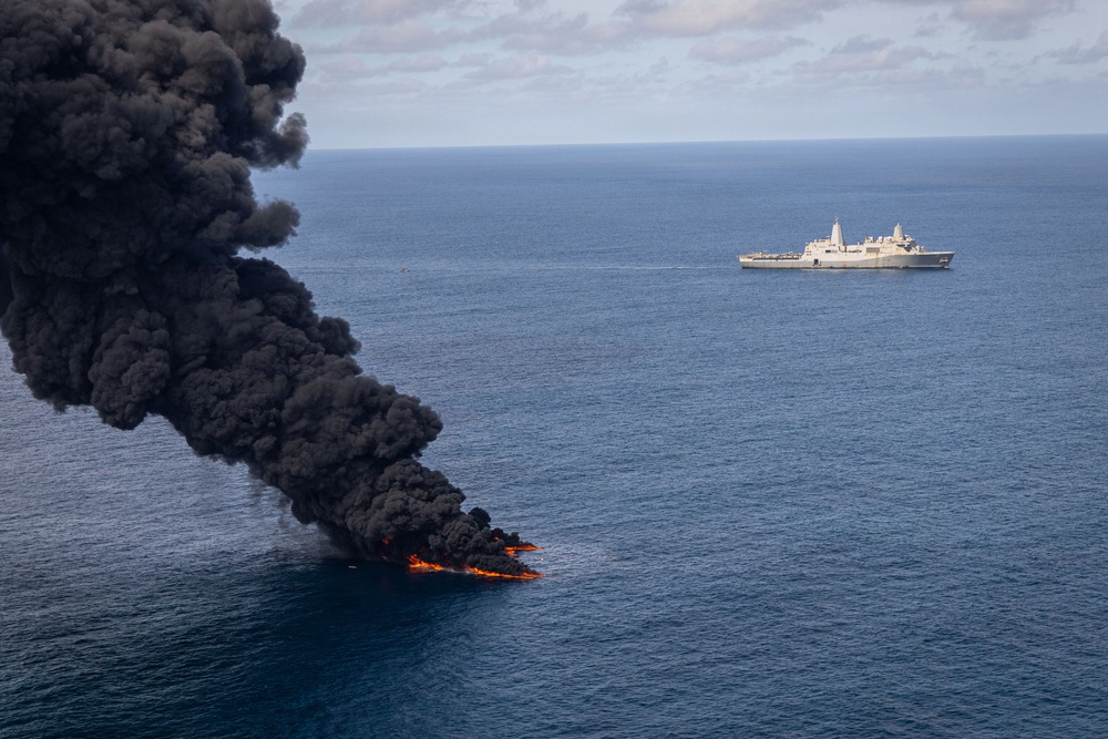 An aerial photograph shows a vessel or object engulfed in intense flames on the open ocean, producing a large column of thick black smoke rising into a partly cloudy sky. In the background, a large gray naval warship — appearing to be a U.S. Navy amphibious transport dock (LPD-class) — is positioned at a safe distance observing the burning vessel. This appears consistent with a controlled destruction or interdiction operation at sea.