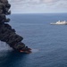 An aerial photograph shows a vessel or object engulfed in intense flames on the open ocean, producing a large column of thick black smoke rising into a partly cloudy sky. In the background, a large gray naval warship — appearing to be a U.S. Navy amphibious transport dock (LPD-class) — is positioned at a safe distance observing the burning vessel. This appears consistent with a controlled destruction or interdiction operation at sea.