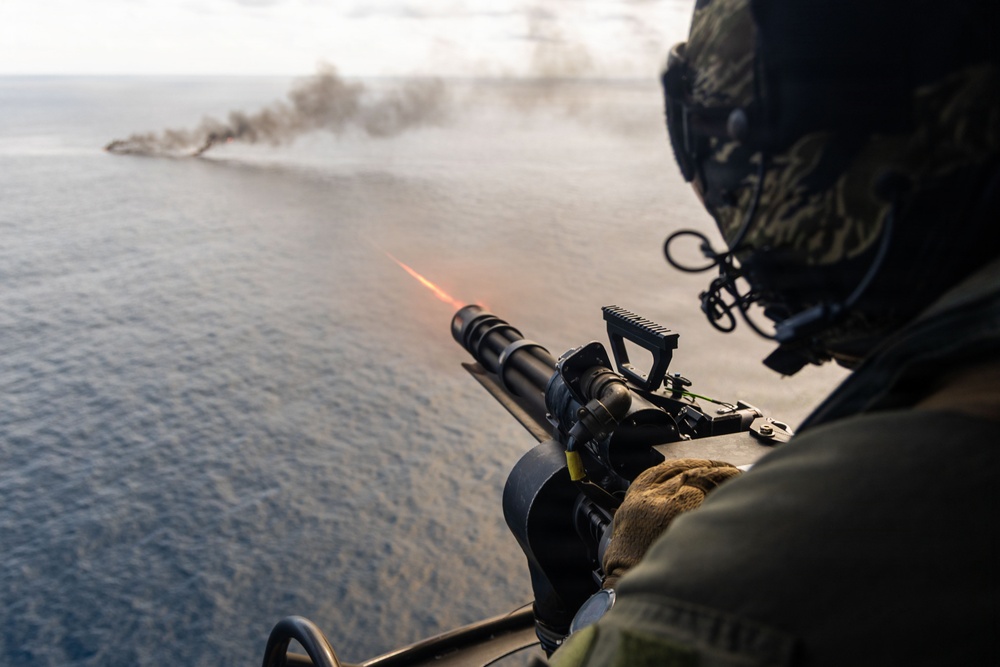 A military aircrew member is firing a door-mounted machine gun from a helicopter over open ocean. A visible tracer round is captured mid-flight, and a smoke plume is visible in the distance on the water surface, indicating an active firing exercise. The shooter is wearing full flight gear including a helmet with communications equipment.