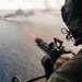 A military aircrew member is firing a door-mounted machine gun from a helicopter over open ocean. A visible tracer round is captured mid-flight, and a smoke plume is visible in the distance on the water surface, indicating an active firing exercise. The shooter is wearing full flight gear including a helmet with communications equipment.