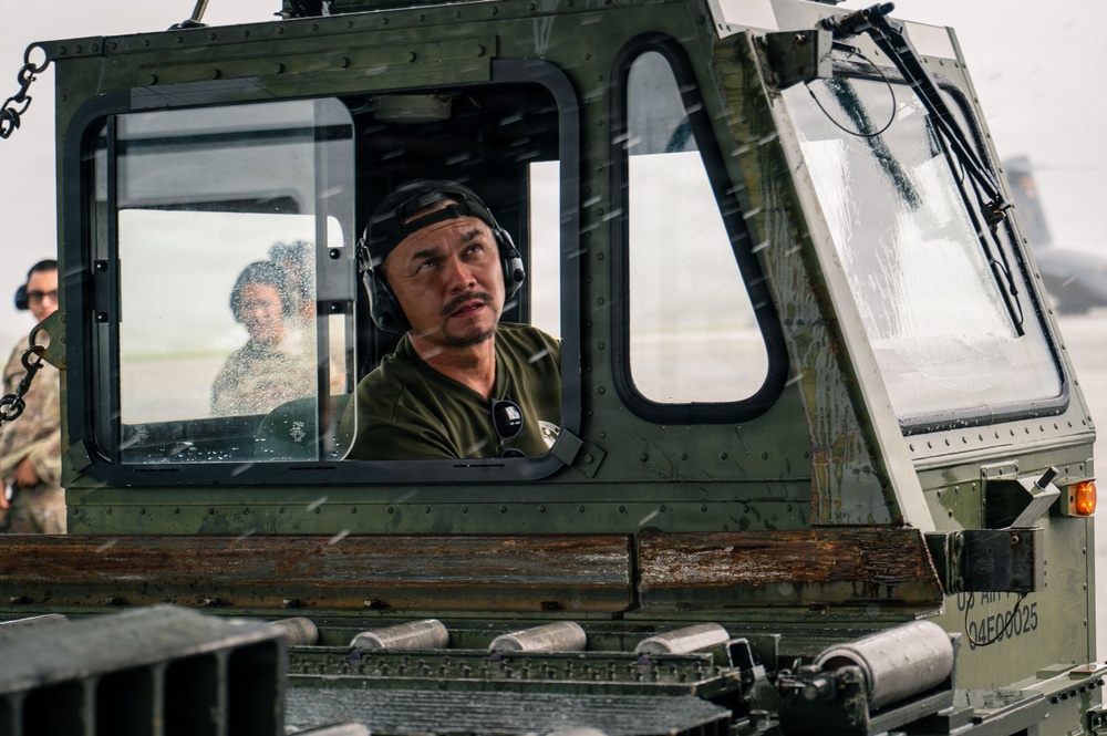A military operator wearing communication headphones and an olive drab shirt is seated inside the cab of a U.S. Army vehicle, looking upward attentively. The vehicle bears a visible U.S. Army identification marking '04E00025' on its exterior. Additional personnel in military attire are visible in the background.