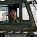 A military operator wearing communication headphones and an olive drab shirt is seated inside the cab of a U.S. Army vehicle, looking upward attentively. The vehicle bears a visible U.S. Army identification marking '04E00025' on its exterior. Additional personnel in military attire are visible in the background.